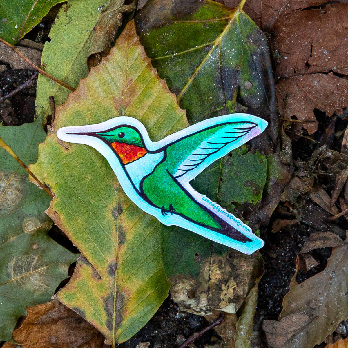A die-cut sticker of a hummingbird with holographic green, blue, and orange details, placed on a background of natural brown and green leaves.