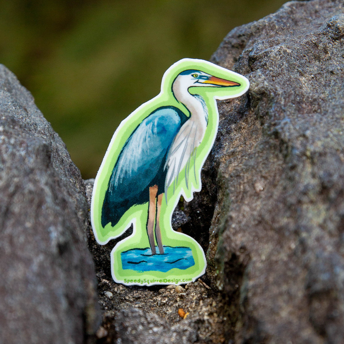 A die-cut sticker of a blue and white heron standing in water, placed on a textured dark rock with a blurred green natural background.