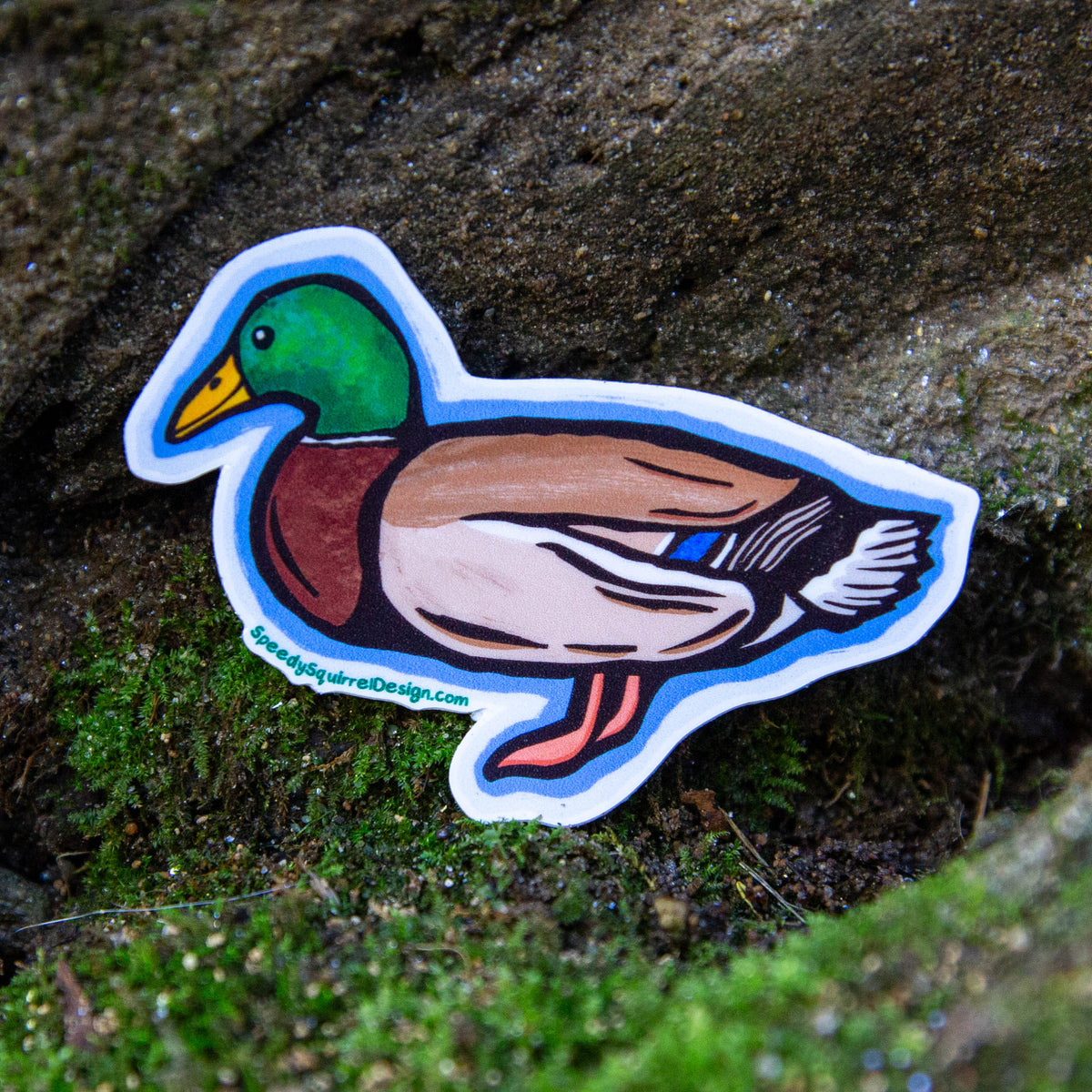 A die-cut vinyl sticker of a mallard duck resting on a mossy, textured tree trunk outdoors. The sticker features a white border and a detailed illustration of a mallard with a green head, yellow beak, reddish-brown chest, brown back, and pinkish legs.
