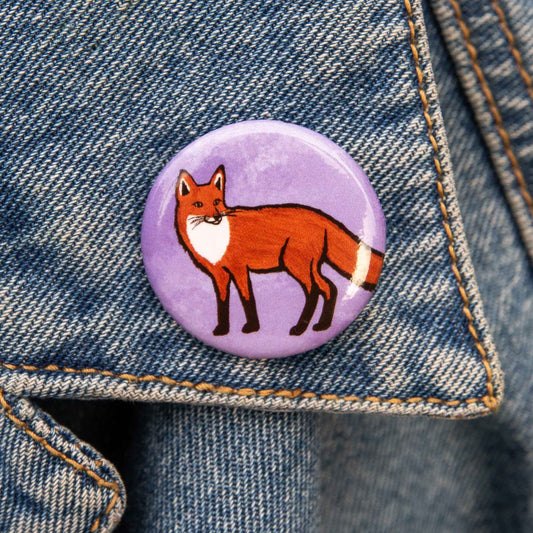 A round pin featuring an illustration of a red fox standing on a textured light purple background, shown against a demin backdrop.