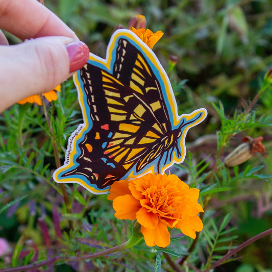 A hand with dark red nail polish holds a die-cut butterfly sticker in front of a green garden background with an orange flower. The sticker features a yellow and black butterfly with blue and orange accents.