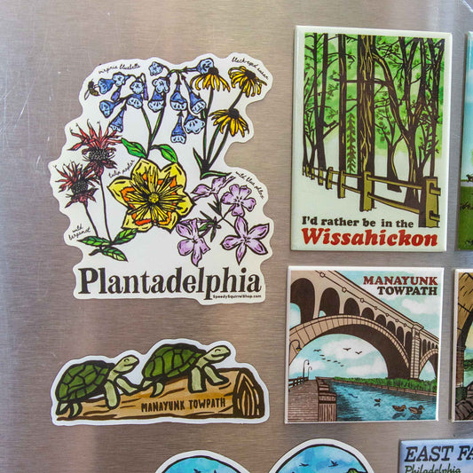 A set of illustrated refrigerator magnets displayed on a brushed metal surface. The main visible magnet reads "Plantadelphia" and features a floral design; other visible magnets include a forest scene labeled "Wissahickon," a towpath bridge scene labeled "Manayunk Towpath," and turtles on a log labeled "Manayunk Towpath.