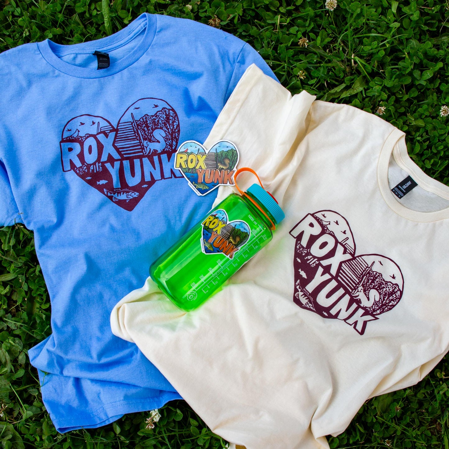 Two t-shirts, a magnet, and sticekr with 'ROX YUNK' branding laying on the grass