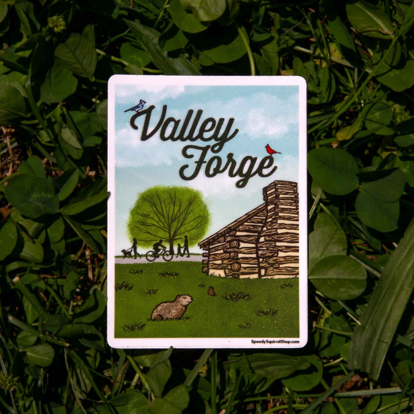 Sticker with 'Valley Forge' design on a grass background. Illustration of a log cabin, tree, and people on a bike and footpath.
