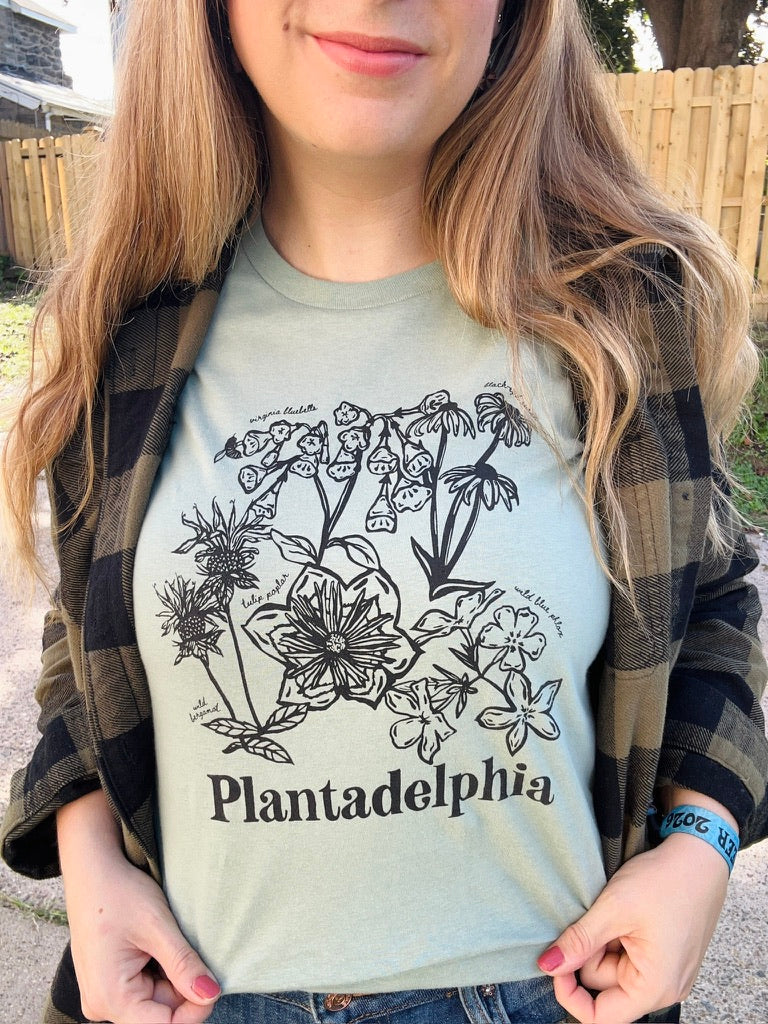 A person wearing a light sage green t-shirt with a black botanical illustration and the text 'Plantadelphia' printed on the front, photographed outdoors.