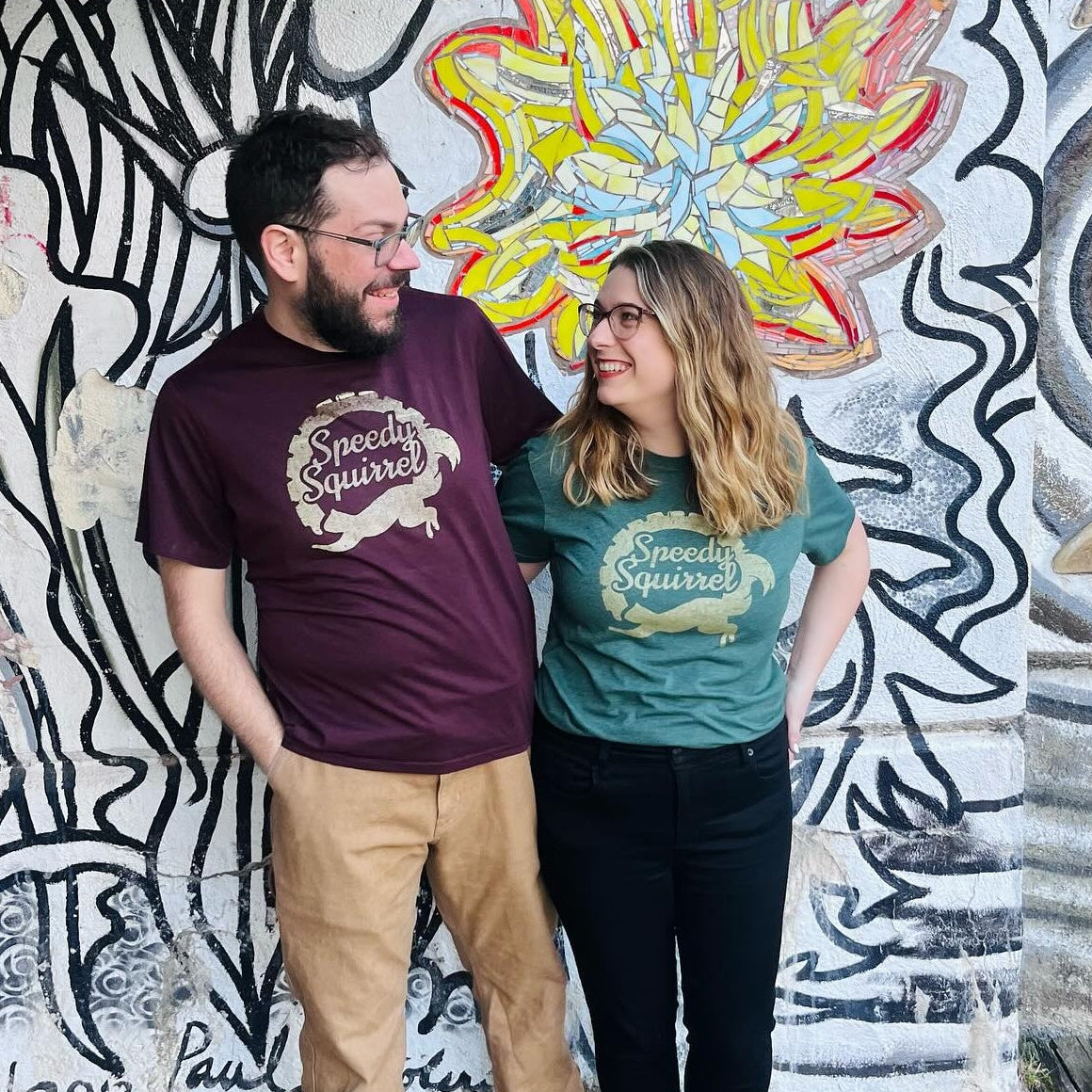 A couple wearing 'Speedy Squirrel' shirts smiling and standing in front of a mosaic-covered wall.