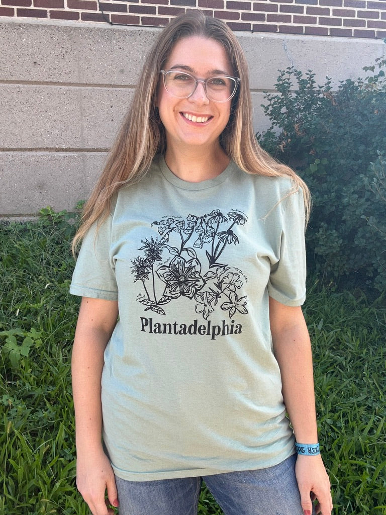 A person wearing a light sage green t-shirt with a black botanical illustration and the text 'Plantadelphia' printed on the front, photographed outdoors in front of a stone wall and greenery.