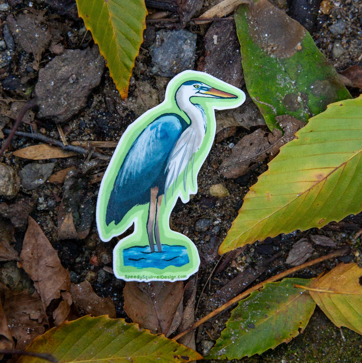 A die-cut sticker of a blue and white heron standing in water, placed on a forest floor among dry leaves, rocks, and green foliage.