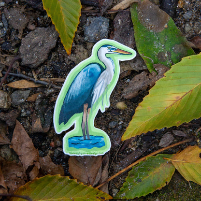 A die-cut sticker of a blue and white heron standing in water, placed on a forest floor among dry leaves, rocks, and green foliage.