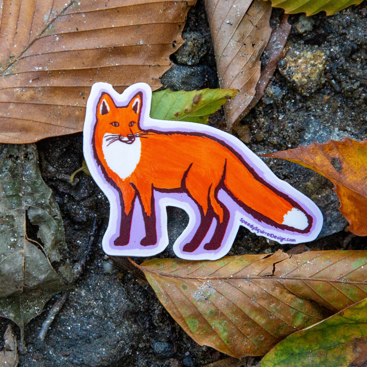 A die-cut sticker of an orange fox with a white chest and tail tip, placed among brown and green autumn leaves.