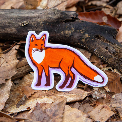 A die-cut sticker of a cartoon fox in orange, white, and brown, placed on a bed of dry brown leaves and a piece of dark wood outdoors.