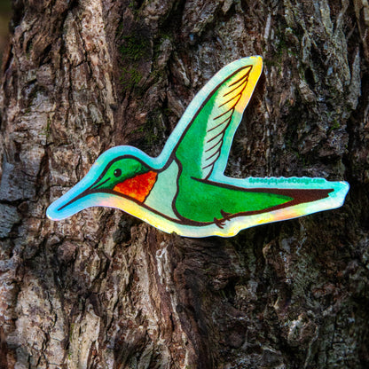 A colorful hummingbird sticker with a holographic iridescent sheen, applied to the side of a textured tree trunk.