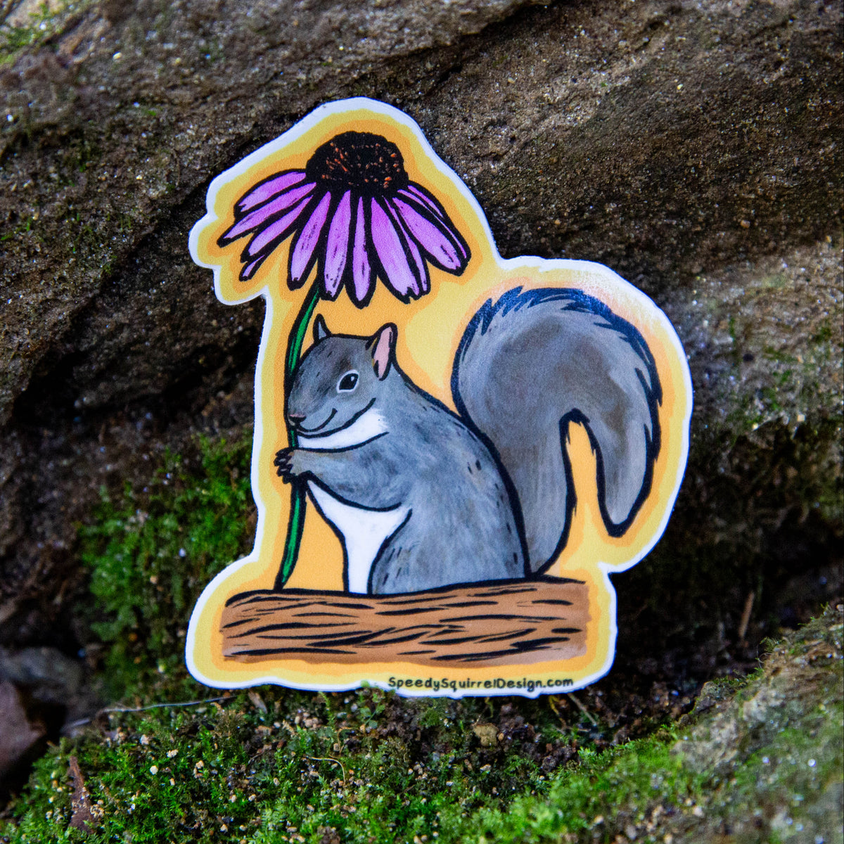 A die-cut sticker featuring a gray squirrel holding a purple coneflower, placed on a mossy rock outdoors. The sticker has a yellow background and is shown against a natural forest backdrop.