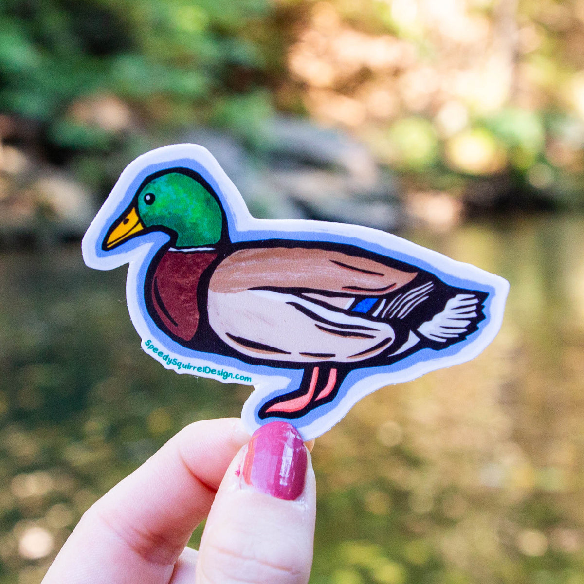 A hand with pink nail polish holds a die-cut sticker of a cartoon mallard duck against a blurred outdoor background of green foliage and water.