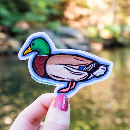 A hand with pink nail polish holds a die-cut sticker of a cartoon mallard duck against a blurred outdoor background of green foliage and water.