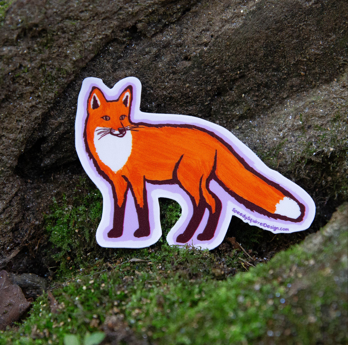A die-cut sticker of an orange fox with white chest and tail tip details, placed on a mossy rock outdoors.