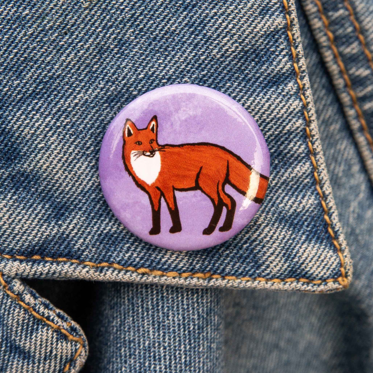 A round pin featuring an illustration of a red fox standing on a textured light purple background, shown against a demin backdrop.
