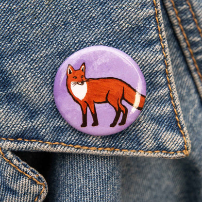 A round pin featuring an illustration of a red fox standing on a textured light purple background, shown against a demin backdrop.