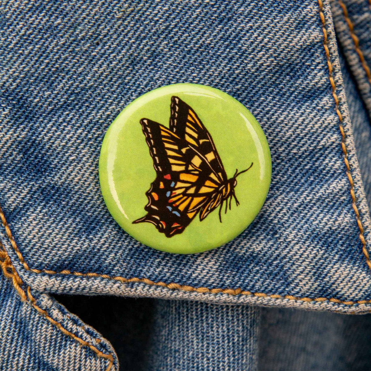 A round pinback button featuring a detailed illustration of a yellow and black butterfly on a light green background, shown pinned to blue denim fabric.