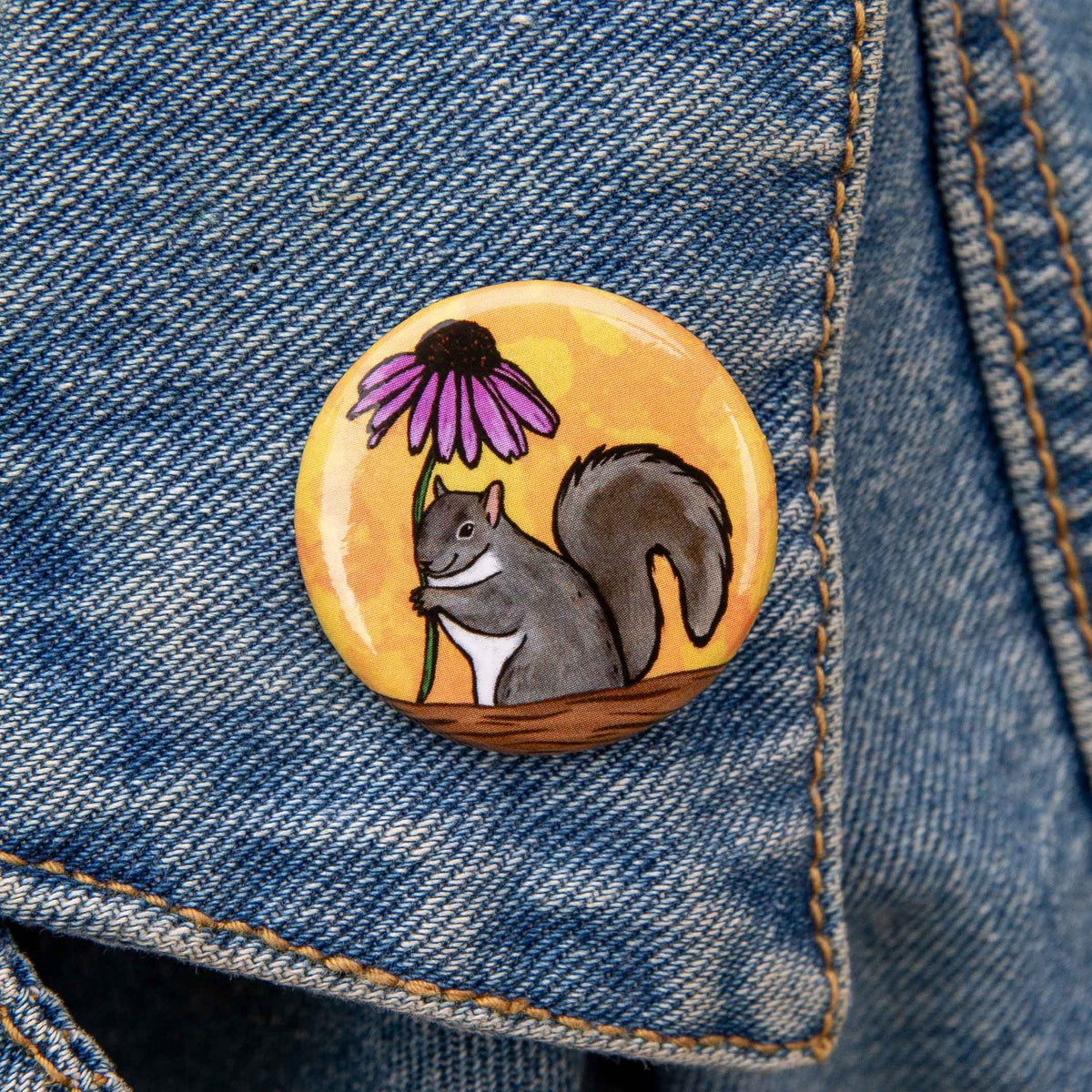 A round pin badge featuring a cartoon gray squirrel holding a purple flower against a yellow-orange background, pinned onto blue denim fabric.