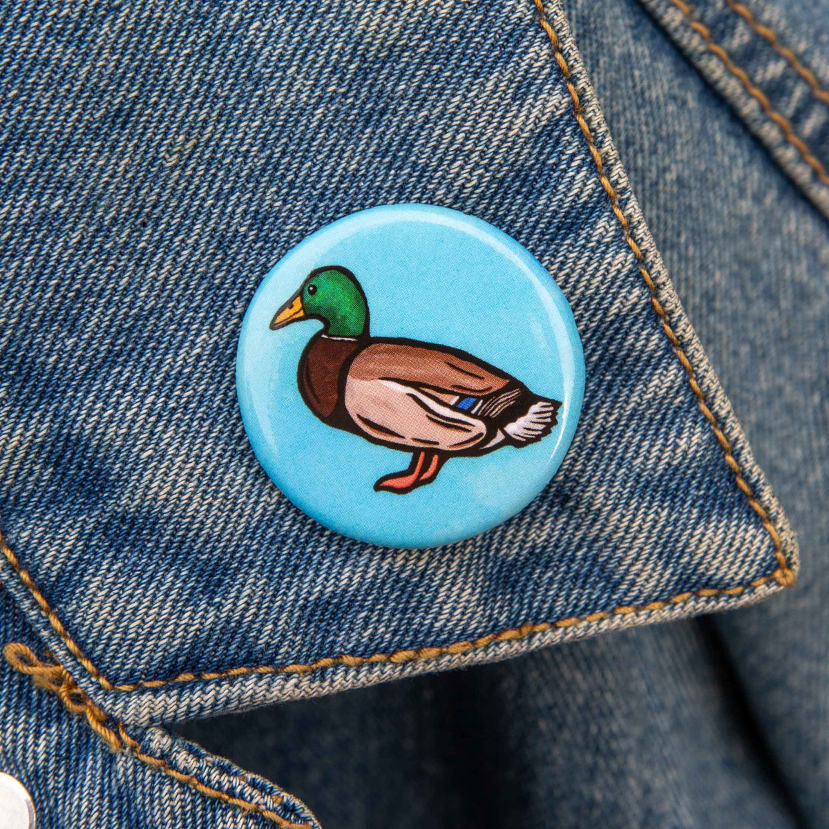 A round pinback button featuring an illustrated mallard duck on a light blue background, pinned onto a blue denim jacket.