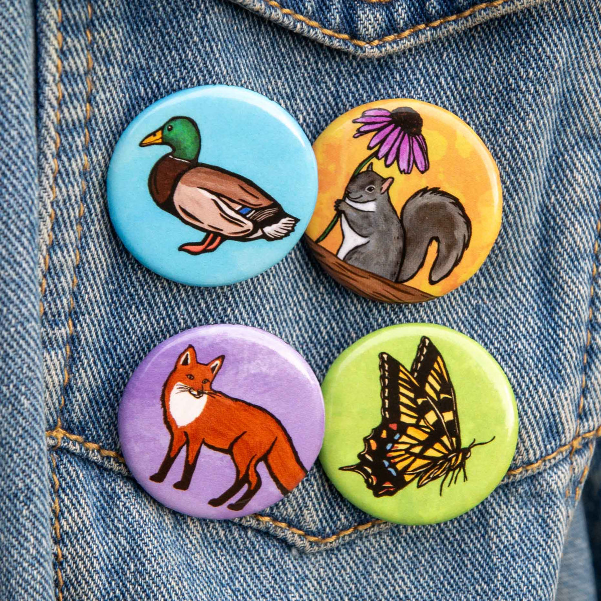 Four round pin-back buttons featuring illustrated nature animals and plants, displayed on a blue denim jacket. The designs include a duck on light blue, a squirrel on yellow, a fox on purple, and a butterfly on green.