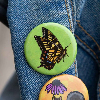 A close-up of a blue denim jacket sleeve with two round pin-back buttons. The top button is light green with a detailed illustration of a black and yellow butterfly.