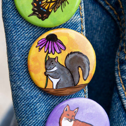 Three round painted pinback buttons attached to a blue denim jacket. The center button shows a gray squirrel holding a purple coneflower on a yellow-orange background, with butterfly and fox designs visible on the top and bottom buttons.