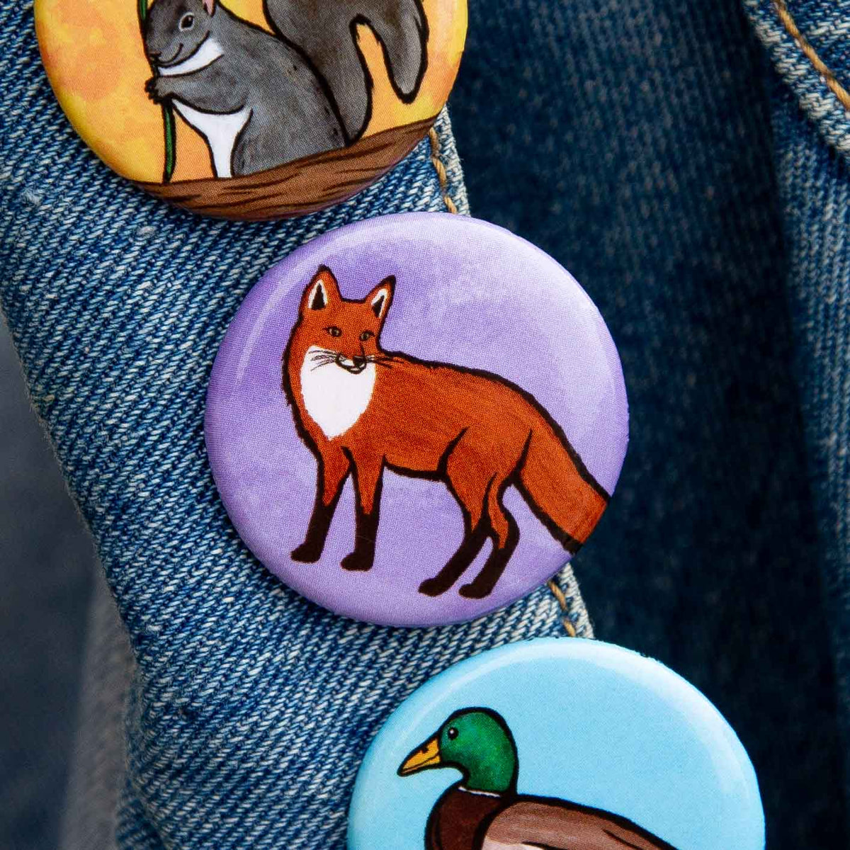 Three round animal-themed pinback buttons attached to a blue denim jacket. The top button shows a gray squirrel on a yellow background, the middle button shows a red fox on a lavender background, and the bottom button shows a mallard duck on a light blue background.