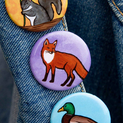 Three round animal-themed pinback buttons attached to a blue denim jacket. The top button shows a gray squirrel on a yellow background, the middle button shows a red fox on a lavender background, and the bottom button shows a mallard duck on a light blue background.