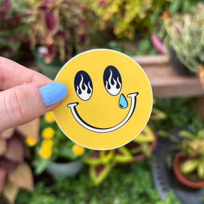 Hand holding a yellow smiley face sticker with a garden in the background. The smiley has a fire design in it's eyes with one tear drop coming down.