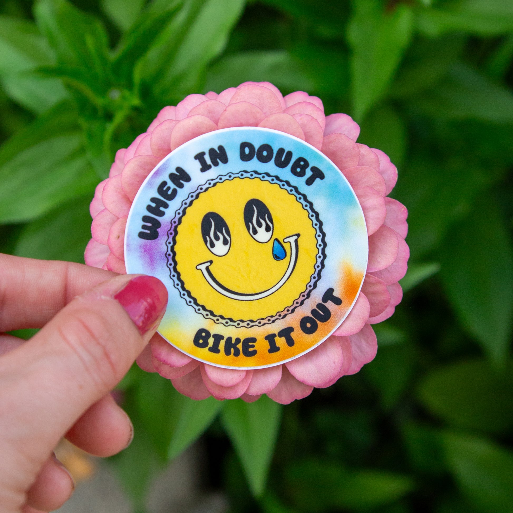 Sticker with smiley face and text 'When in doubt, bike it out' held by a hand against a green leafy background