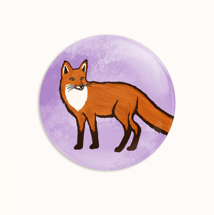 A round pin badge featuring an illustration of a red fox standing on a textured light purple background, shown against a plain off-white backdrop.
