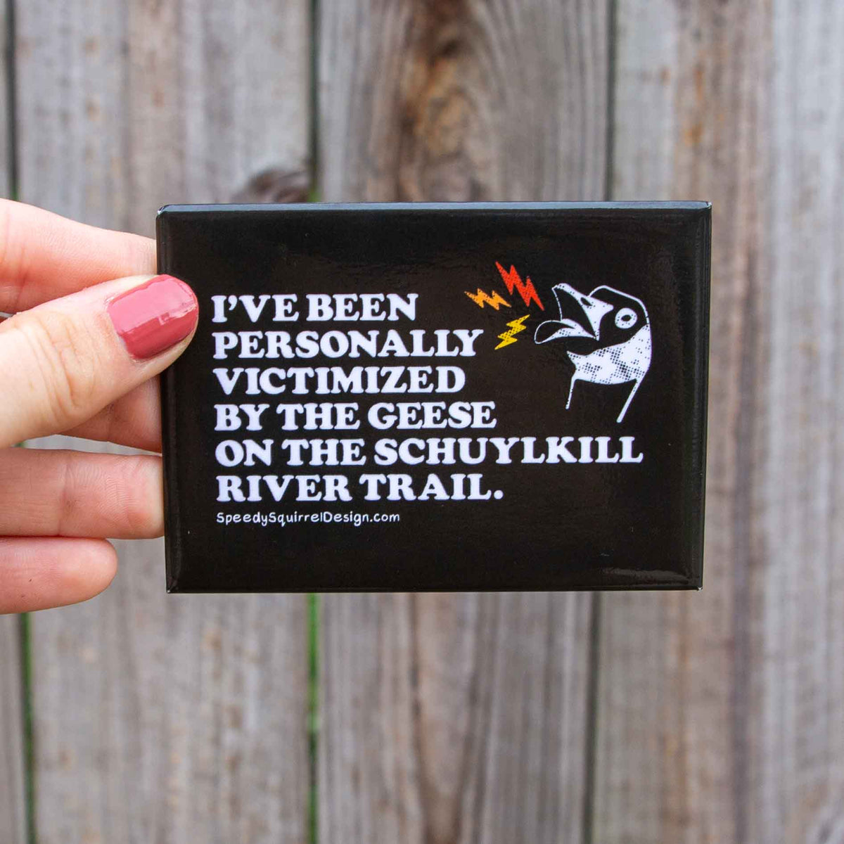A black rectangular fridge magnet with text 'I've been personally victimized by the geese in the Schuylkill River Trail, featuring a goose illustration and small red and yellow lightning accents, in front of a wooden fence.