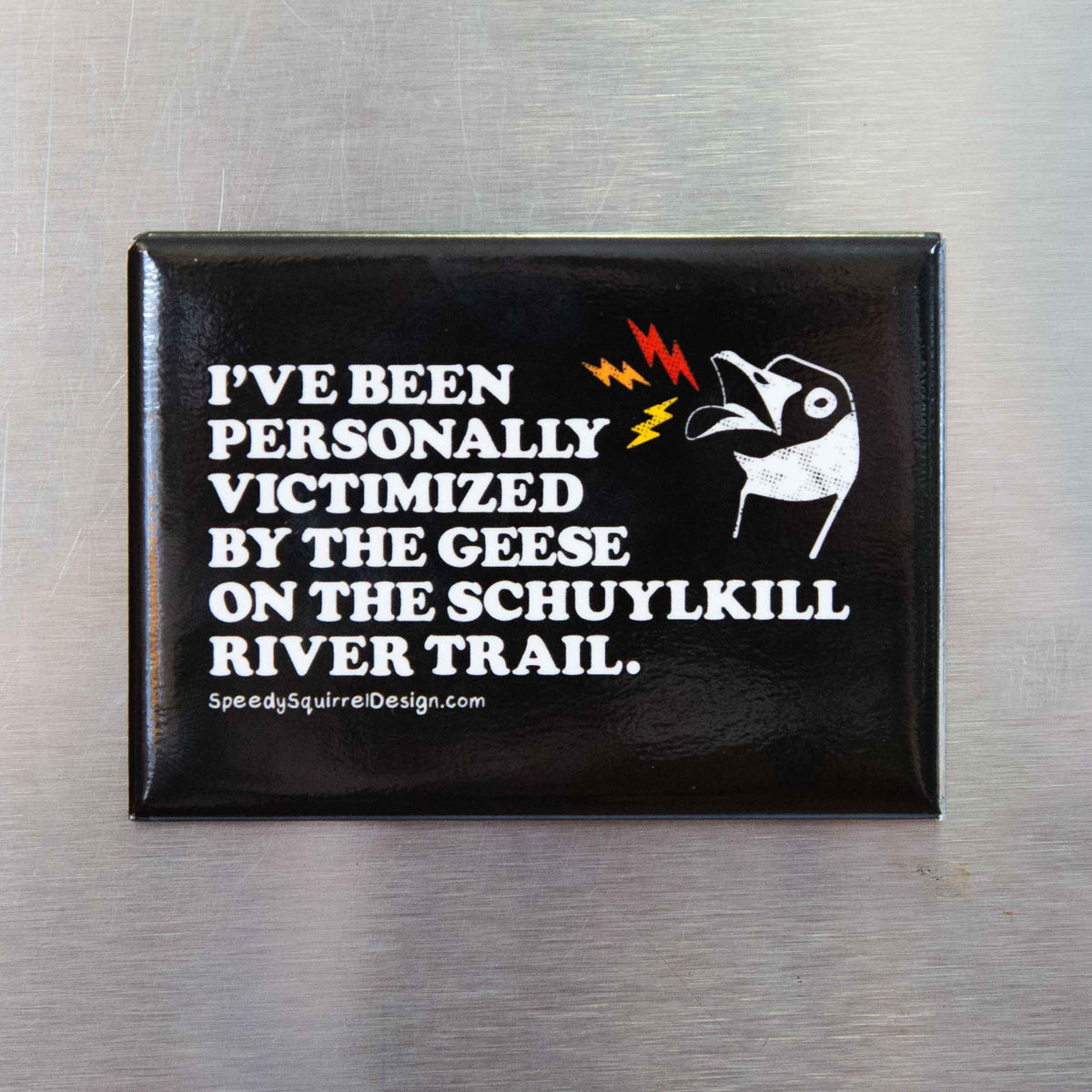 A black rectangular fridge magnet with text 'I've been personally victimized by the geese in the Schuylkill River Trail, featuring a goose illustration and small red and yellow lightning accents, placed on a brushed metal surface.