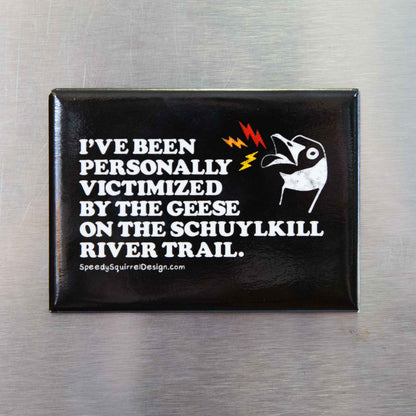 A black rectangular fridge magnet with text 'I've been personally victimized by the geese in the Schuylkill River Trail, featuring a goose illustration and small red and yellow lightning accents, placed on a brushed metal surface.