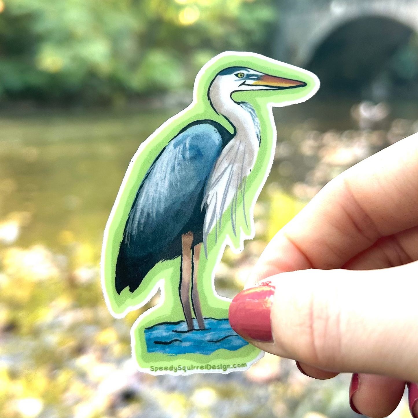 A die-cut sticker featuring a painted blue-gray heron standing in water, held by a hand against a blurred outdoor background of a stream and greenery.