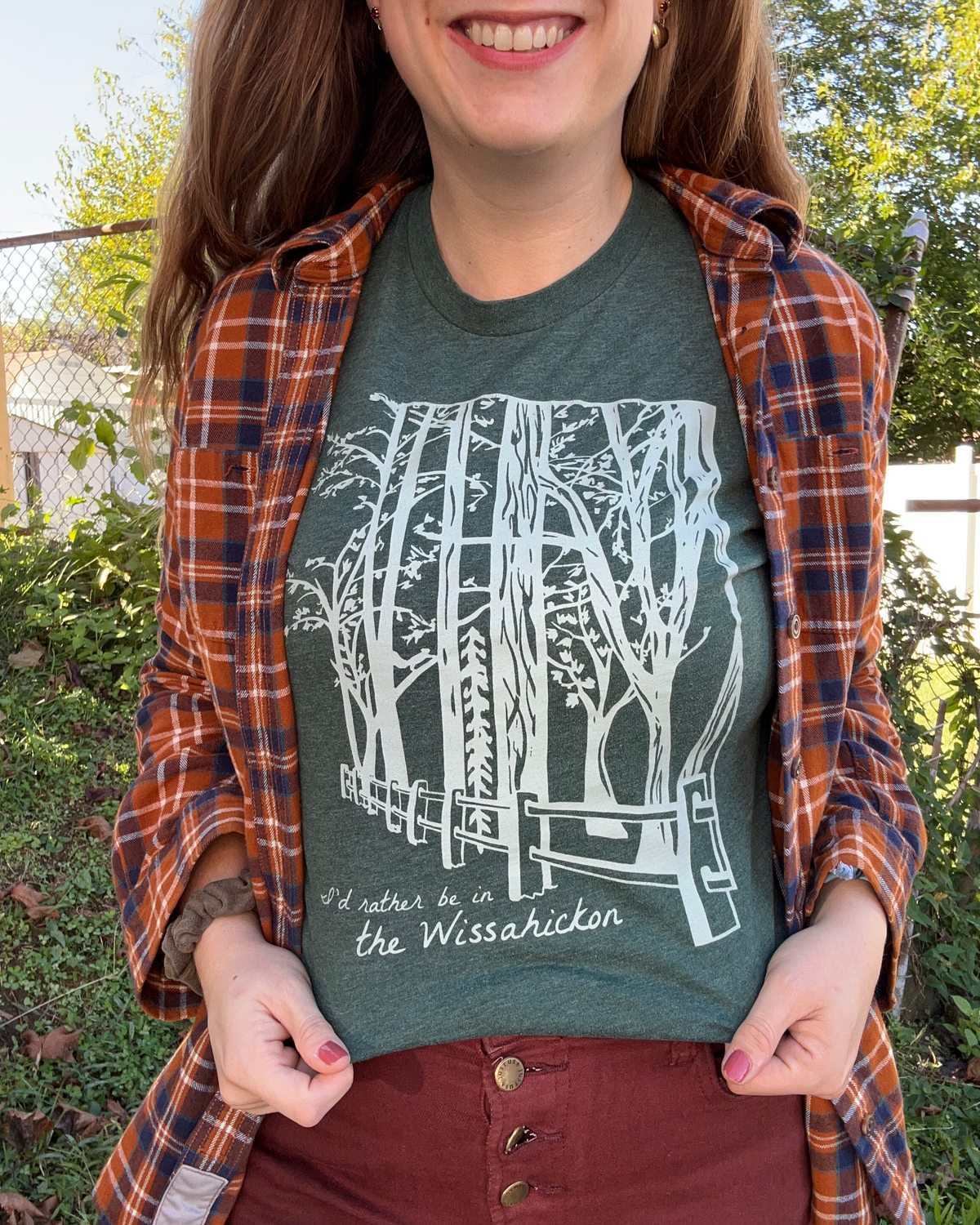 Young women wearing a dark green t-shirt and orange flannel with a light green fence and forest with text 'I'd rther be in the Wissahickon' while standing outdoors and smiling.