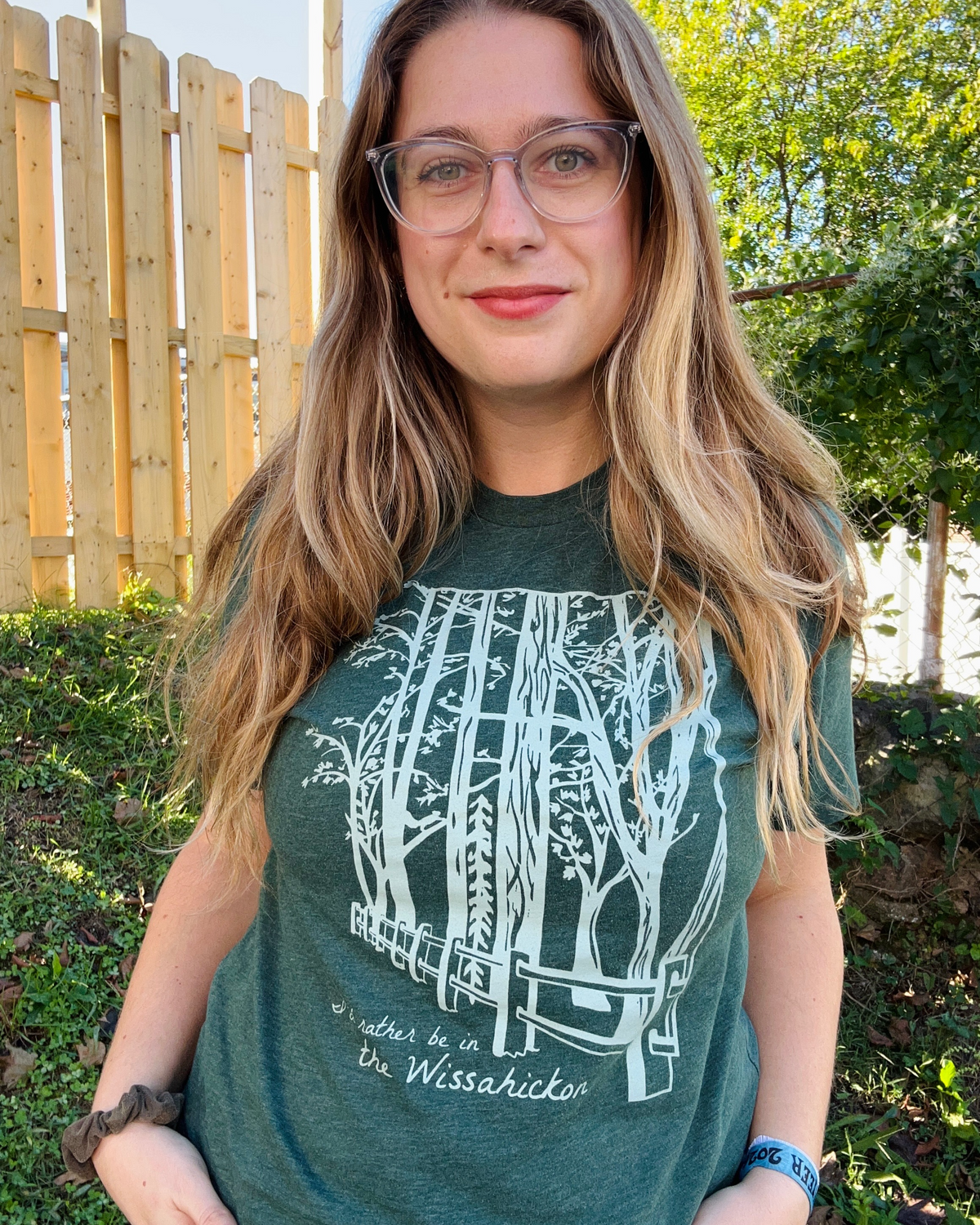 Young women wearing a dark green t-shirt with a light green fence and forest with text 'I'd rther be in the Wissahickon' while standing outdoors and smiling.