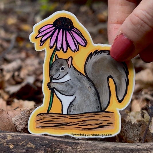 A hand with pink nail polish holds a die-cut sticker featuring a gray squirrel holding a pink coneflower against a yellow background, with a forest floor backdrop.