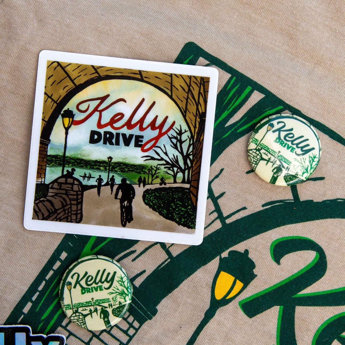 A square sticker with a white border featuring an illustrated scene under an archway with the text 'Kelly Drive,' shown on a beige fabric surface alongside two matching round pins.