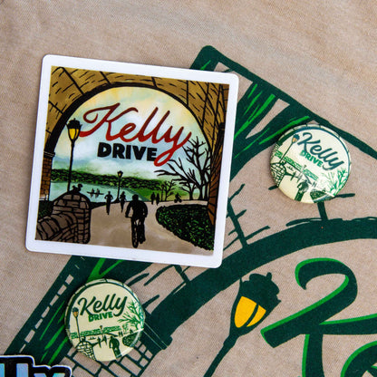 A square sticker with a white border featuring an illustrated scene under an archway with the text 'Kelly Drive,' shown on a beige fabric surface alongside two matching round pins.