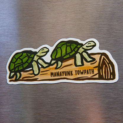  two turtles resting on a log with "Manayunk Towpath" written across it on a metallic background. 