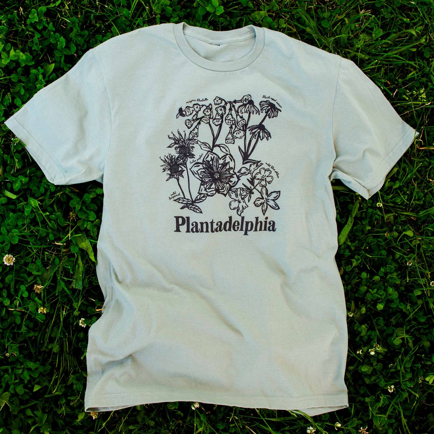 Sage green t-shirt with floral design and 'Plantadelphia' text on a grassy background