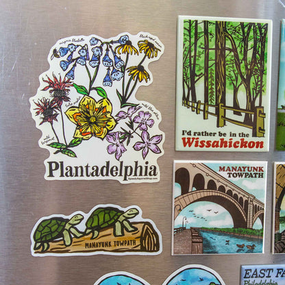 A set of illustrated refrigerator magnets displayed on a brushed metal surface. The main visible magnet reads "Plantadelphia" and features a floral design; other visible magnets include a forest scene labeled "Wissahickon," a towpath bridge scene labeled "Manayunk Towpath," and turtles on a log labeled "Manayunk Towpath.