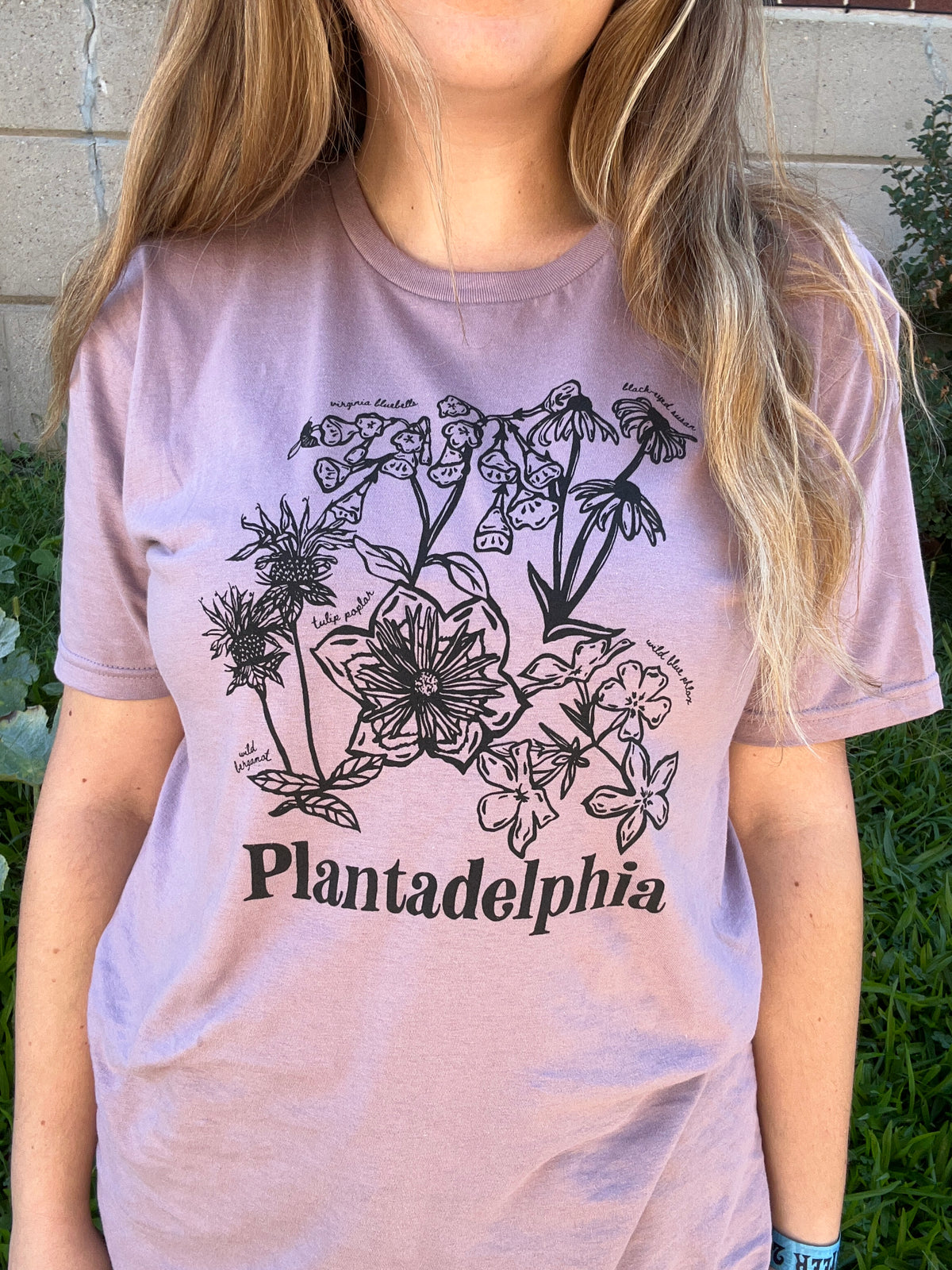 A person wearing a light mauve-pink t-shirt with a black botanical illustration and the text 'Plantadelphia' printed on the front, photographed outdoors against a stone wall and greenery.