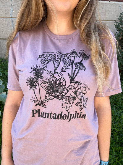 A person wearing a light mauve-pink t-shirt with a black botanical illustration and the text 'Plantadelphia' printed on the front, photographed outdoors against a stone wall and greenery.