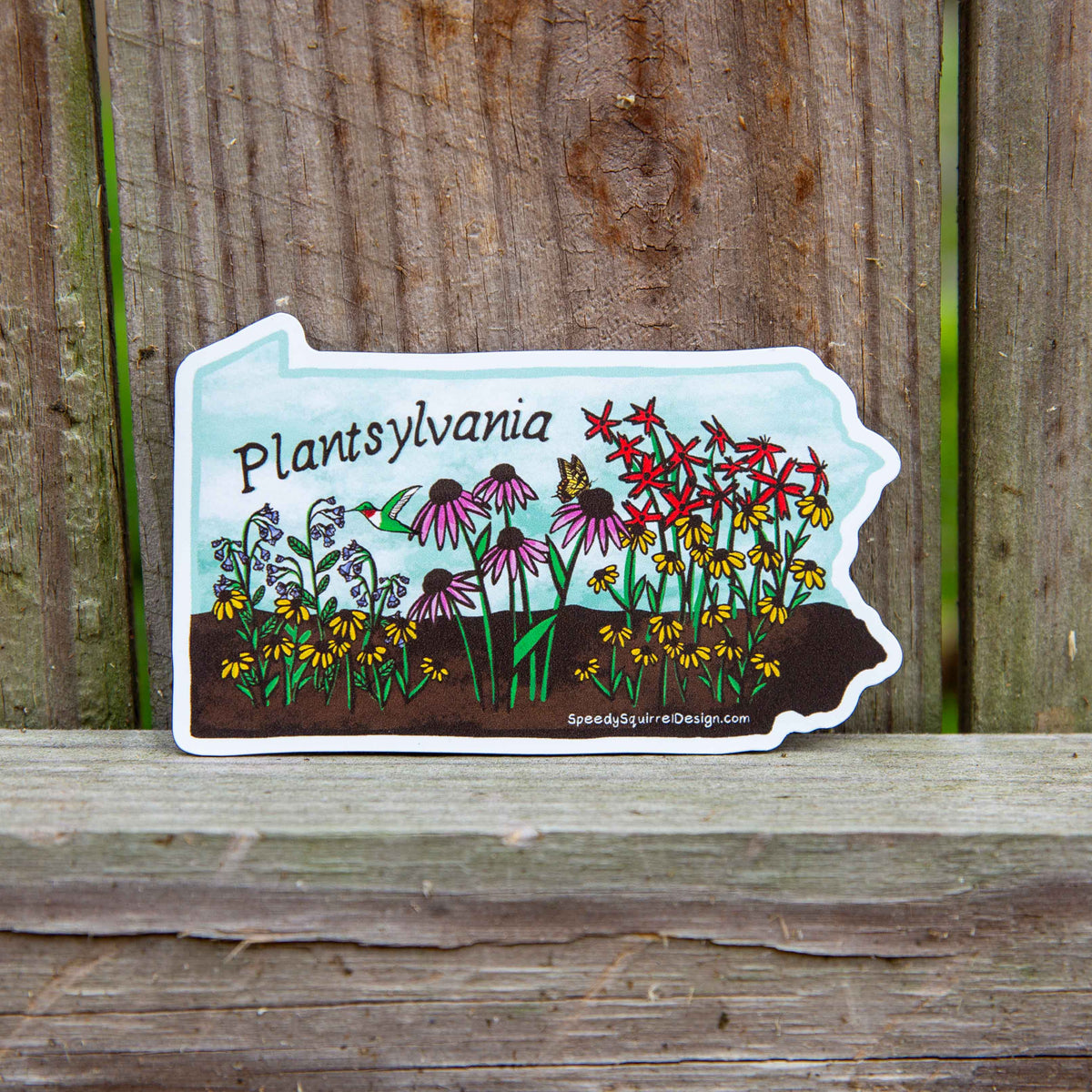 A die-cut sticker shaped like the state of Pennsylvania, featuring a colorful illustration of wildflowers, a hummingbird, and the text 'Pennsylvania' and SpeedySquirrelDesign.com', placed on a weathered wooden surface.