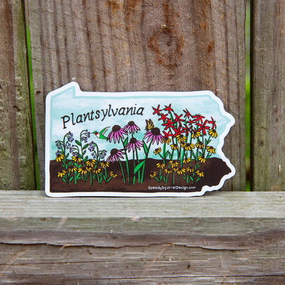 A die-cut sticker shaped like the state of Pennsylvania, featuring a colorful illustration of wildflowers, a hummingbird, and the text 'Pennsylvania' and SpeedySquirrelDesign.com', placed on a weathered wooden surface.