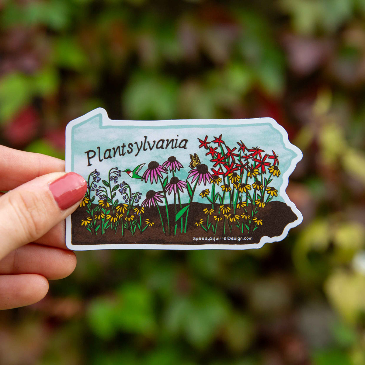 A hand with pink nail polish holds a die-cut sticker shaped like the state of Pennsylvania, featuring an illustration of wildflowers and the text 'Plantsylvania' against a blurred green outdoor background.